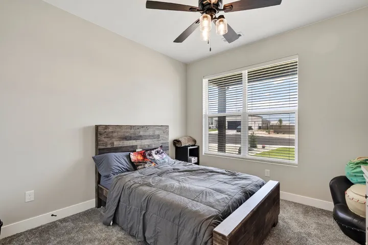 Textured carpet flooring, window with horizontal blinds, overhead ceiling fan with light fixture, and light-toned wall paint