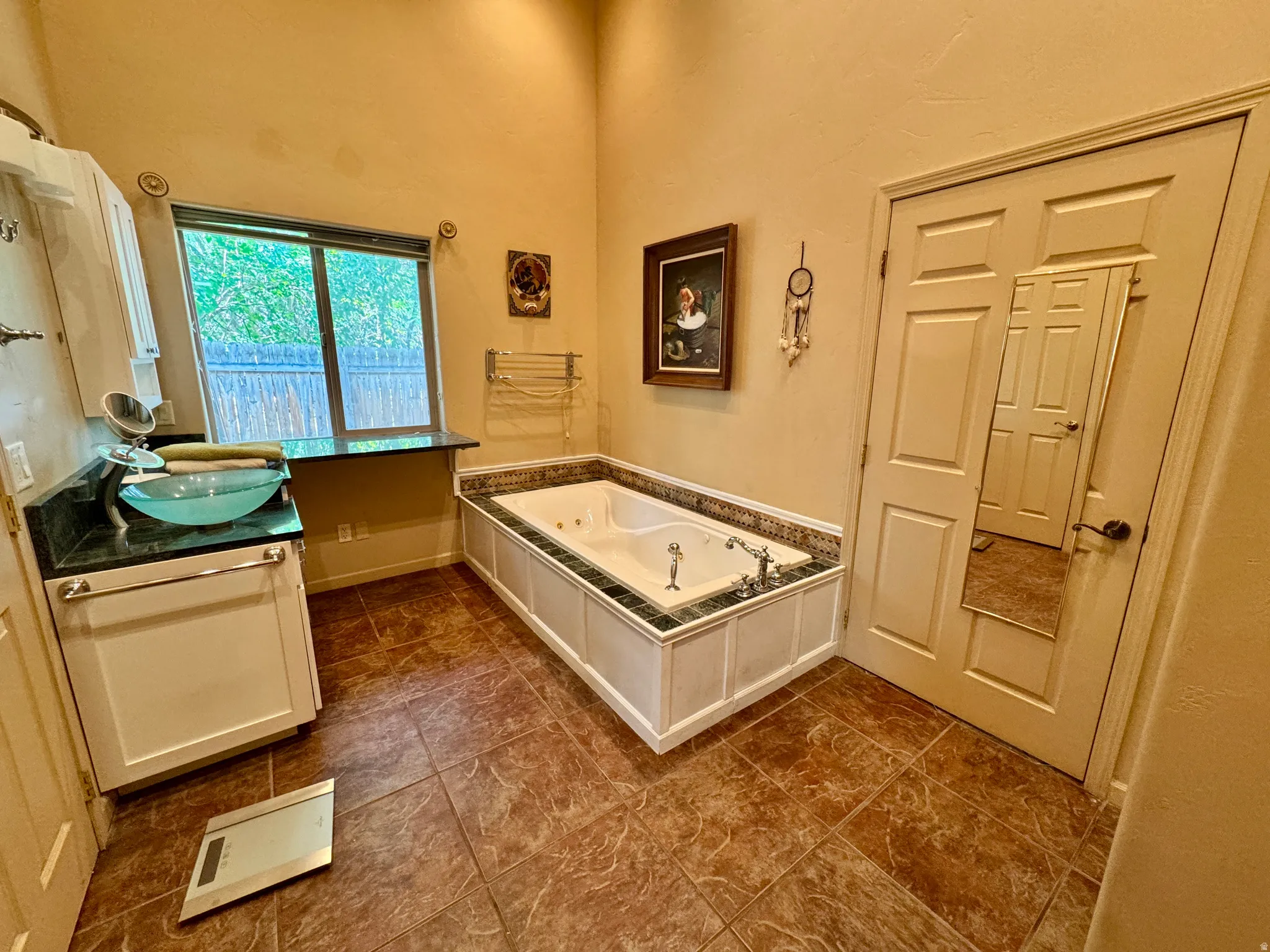 Jetted soaking tub with tile surround, single vanity with vessel sink and dark countertop, and tiled flooring