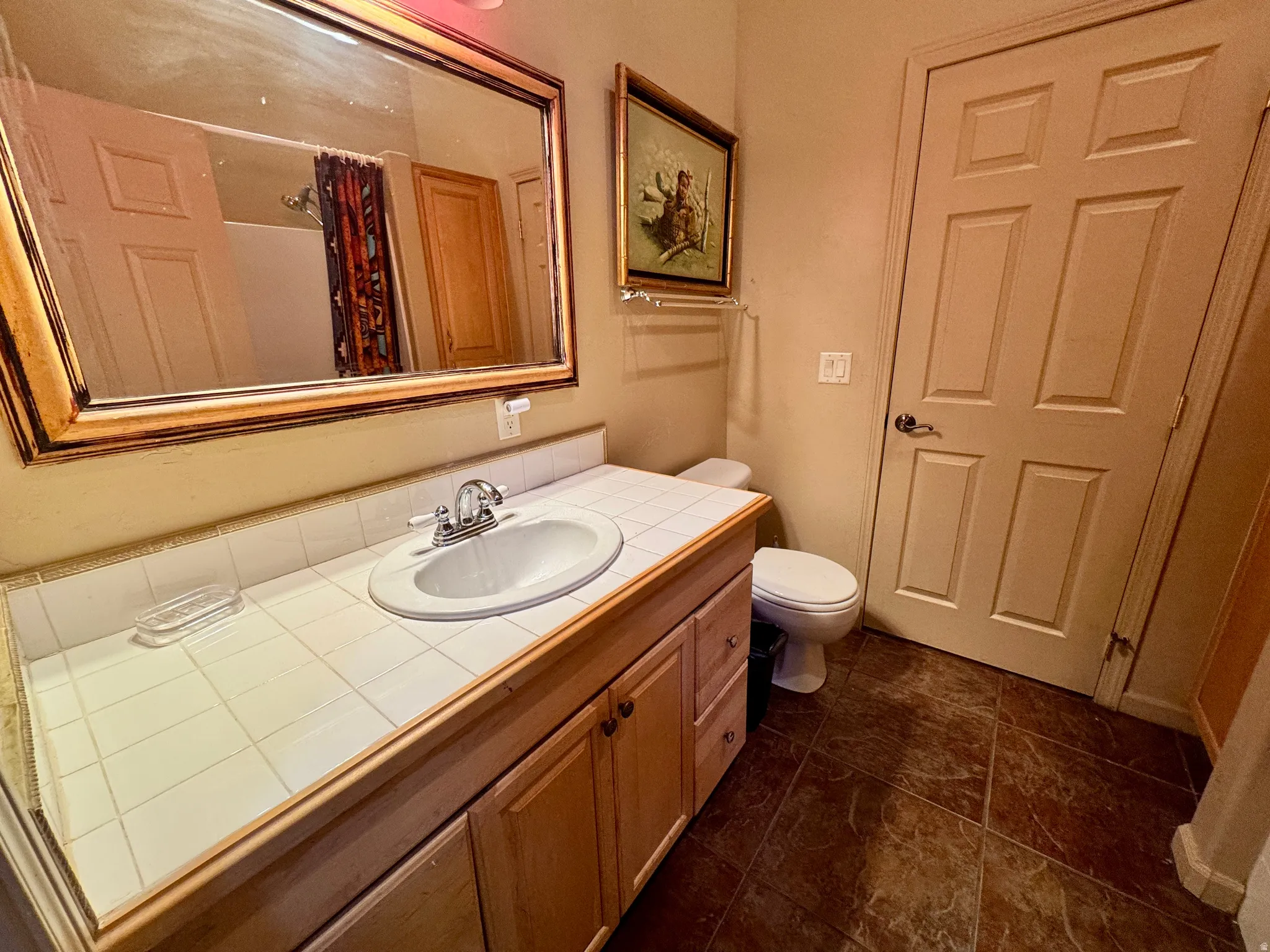 Vanity with integrated sink, tile countertop, wood-finish cabinetry, and framed mirror