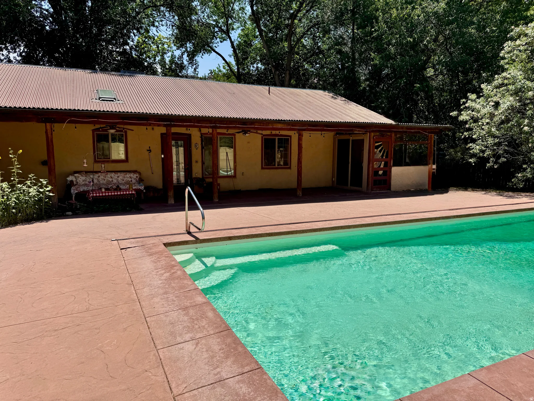 Outdoor swimming pool with integrated steps, expansive concrete decking, and covered patio featuring wood support posts and a metal roof