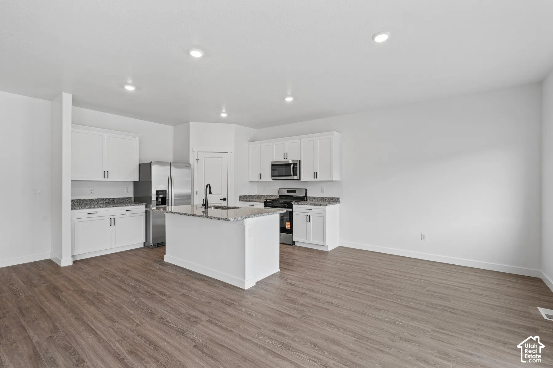 Open-concept kitchen featuring white cabinetry, granite countertops, stainless steel appliances, a kitchen island with a black faucet, and wood-finish flooring
