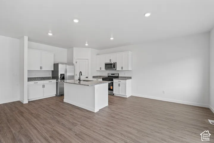 Open-concept kitchen featuring white cabinetry, granite countertops, stainless steel appliances, a kitchen island with a black faucet, and wood-finish flooring