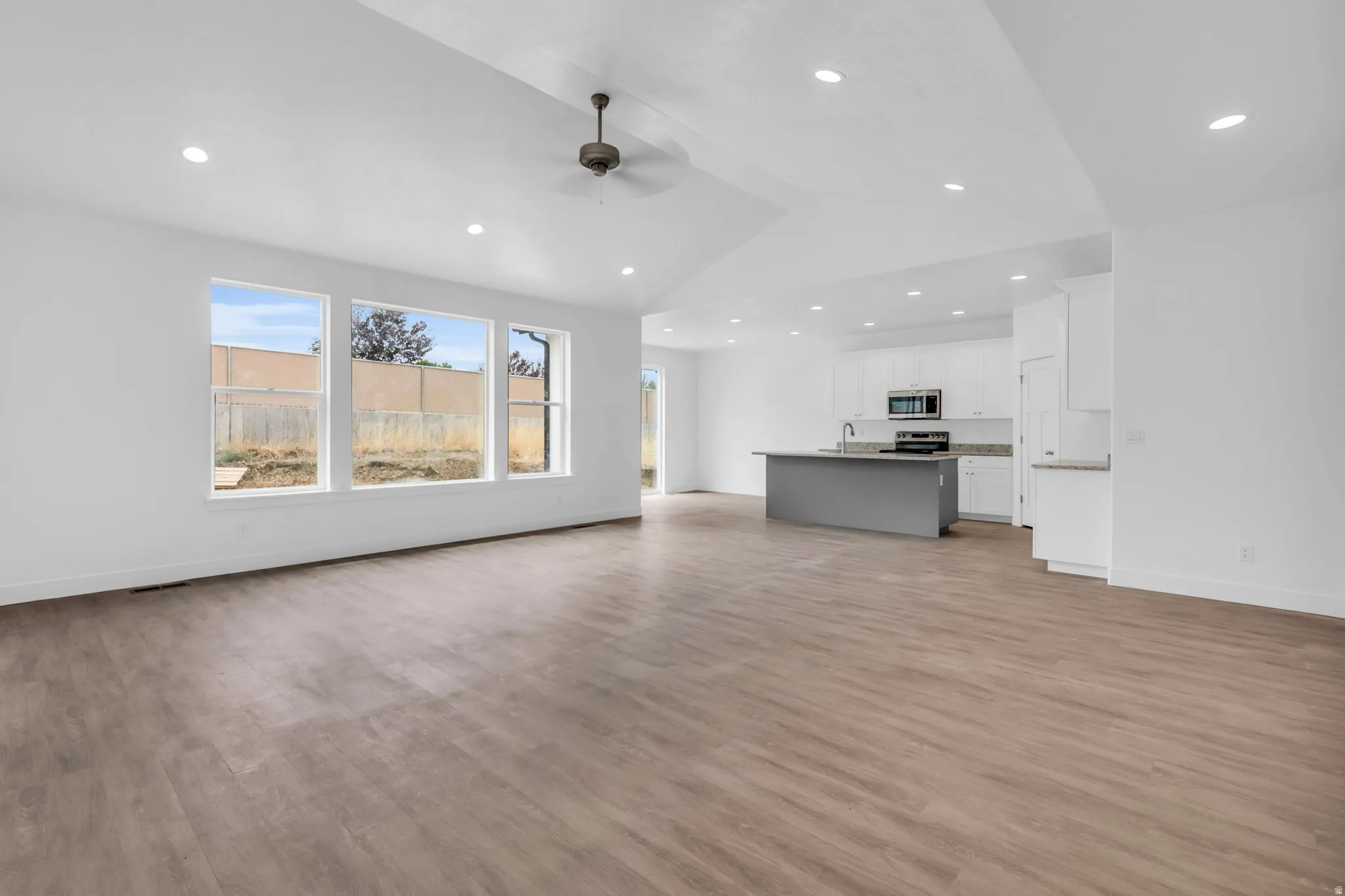 Vaulted ceiling with recessed lighting and a ceiling fan, wood-finish flooring, a kitchen island with a sink, white cabinetry, and stainless steel appliances