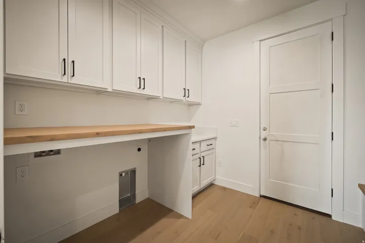 Main floor laundry room with exit to garage features white shaker cabinetry with matte black hardware, wood-finish countertops, wood-finish flooring, and a paneled interior door