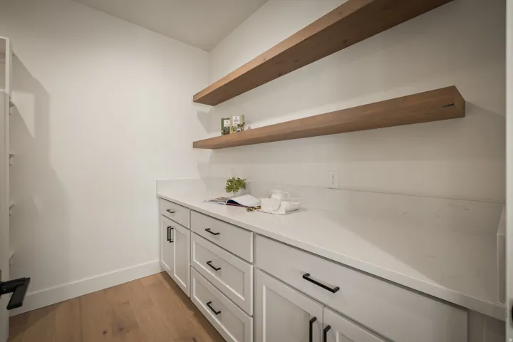Butlers pantry features white cabinetry with black hardware, light-toned countertop, wood-finish flooring, and two floating wood shelves