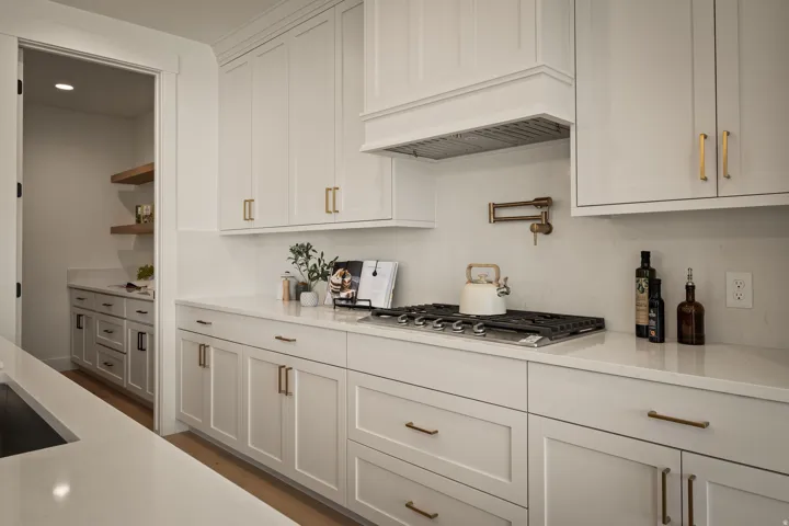 Chef's kitchen featuring white shaker-style cabinetry with brushed gold hardware, quartz countertops, a gas cooktop with stainless steel range hood, and a butler's pantry with pass-through access