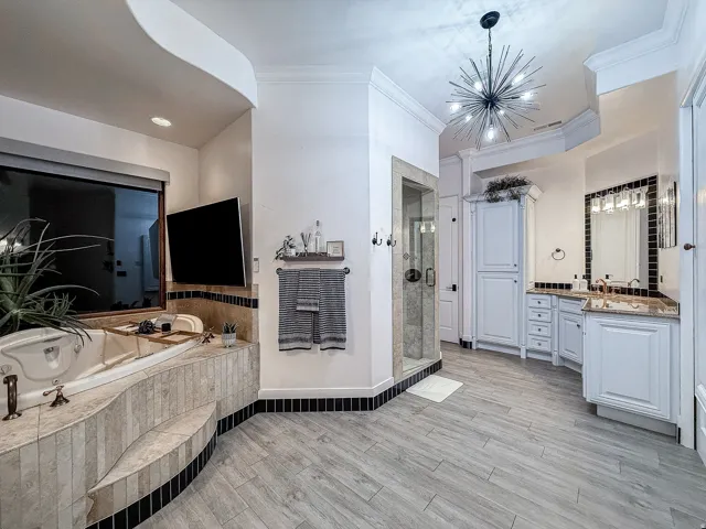 Built-in jetted tub with tiled surround, walk-in glass shower, double vanity with stone countertop, wood-finish flooring, and a contemporary chandelier