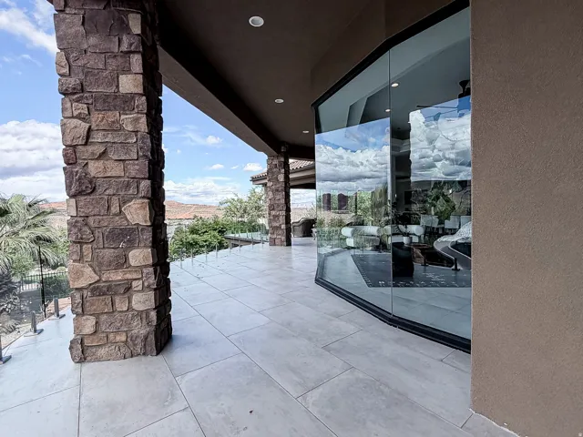 Expansive outdoor patio featuring light-toned tile flooring, stone-clad columns, and a glass safety railing