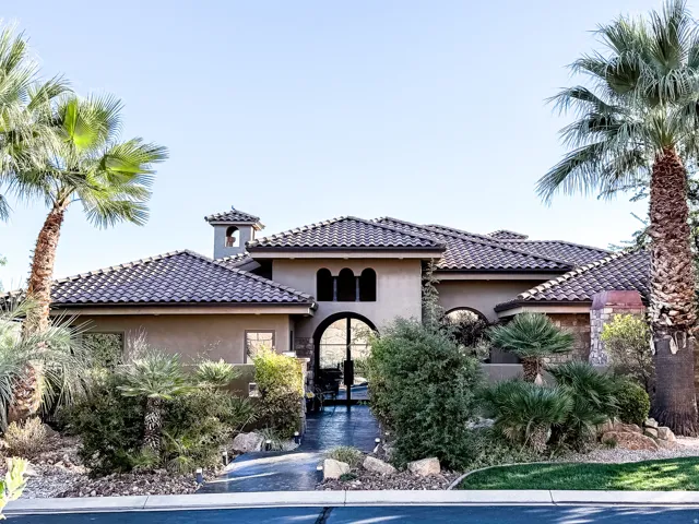 Mediterranean-inspired residence featuring a stucco exterior with clay tile roofing, arched entryway with dark wood detailing, and a circular driveway framed by mature palm trees and desert landscaping