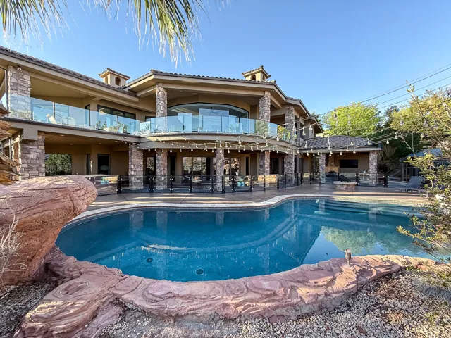 Expansive backyard featuring a custom-shaped swimming pool, integrated rock formations, a covered outdoor kitchen area, and a stone-clad exterior with a second-story glass-paneled balcony