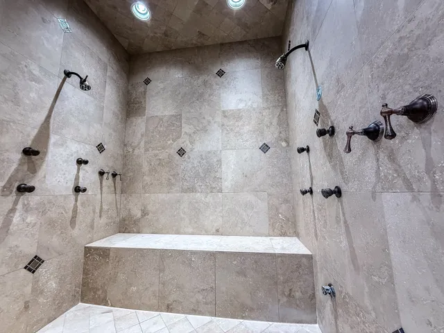 Expansive shower featuring stone-finish wall and floor tiles, dual shower heads, multiple body sprays, a built-in bench, and recessed lighting