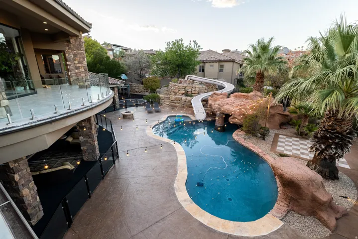 Curved swimming pool with rock features and an integrated slide, expansive patio with string lighting, fire pit, and a full-height stone-clad exterior with a glass railing balcony