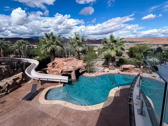 Freeform swimming pool with a rock grotto, integrated water slide, stamped concrete patio, and a glass railing