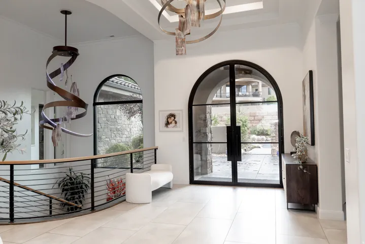 Arched entry door with black framing, tile flooring, curved railing with horizontal metal balusters, and a contemporary chandelier