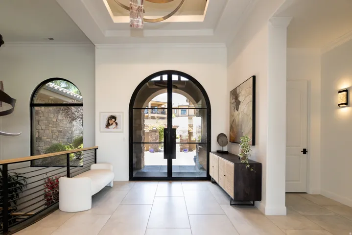 Arched iron-frame double entry doors, coffered ceiling with a chandelier, light-toned tile flooring, and a contemporary railing system with horizontal balusters