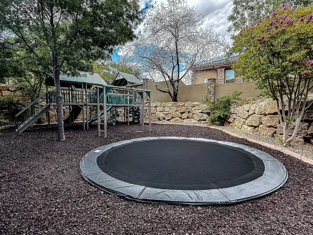 Expansive backyard featuring a large in-ground trampoline, a multi-level playset with slides, and mature trees