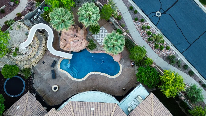 Curved swimming pool with rock grotto and water slide, surrounded by a concrete patio and mature landscaping
