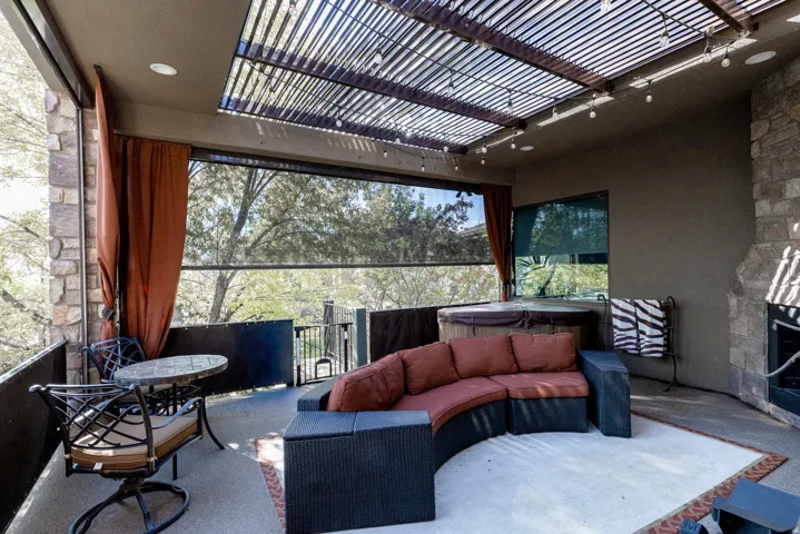 Covered outdoor living space featuring a slatted pergola, stone-clad fireplace, built-in hot tub, and retractable window shades