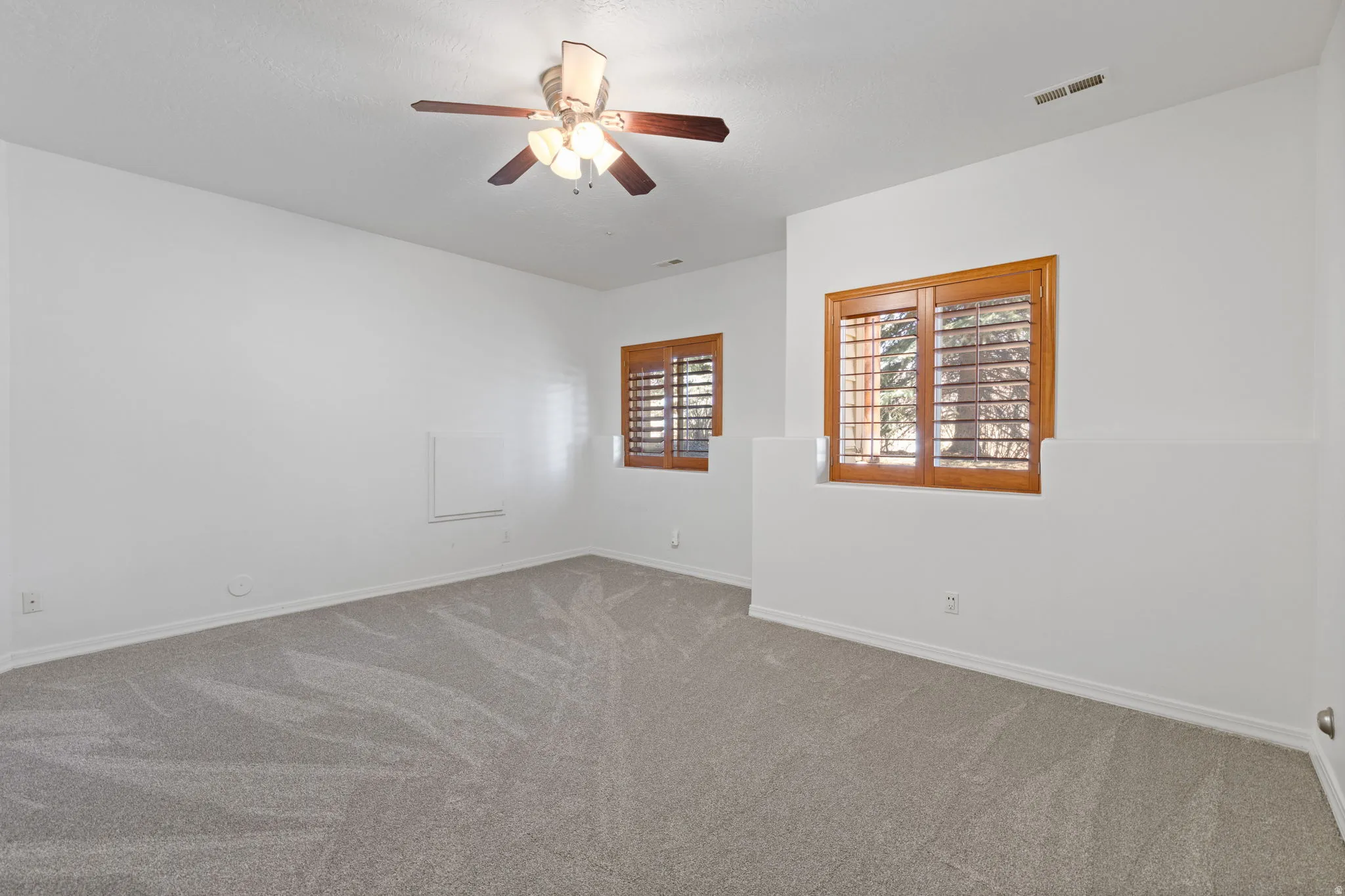 Spacious room featuring light gray carpeting, crisp white walls, two windows with wood plantation shutters, and a ceiling fan with integrated lighting