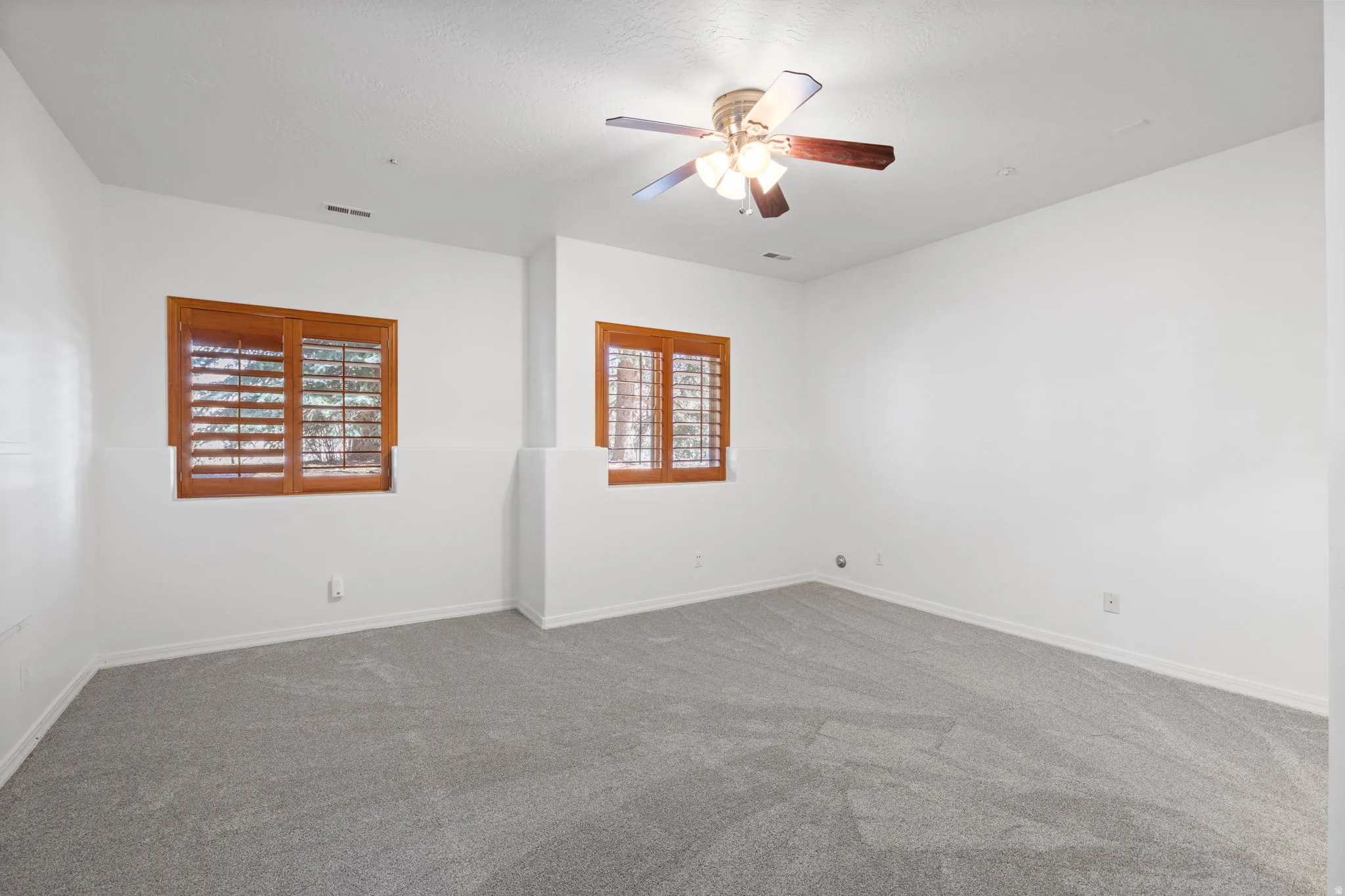 Spacious room featuring light grey carpeting, white walls, two windows with wooden plantation shutters, and a ceiling fan with light fixture