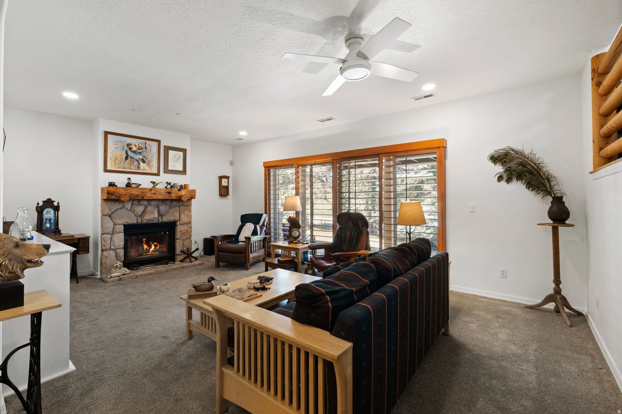 Living area featuring a stone fireplace with a wood mantle, large windows with wood blinds, a ceiling fan, and recessed lighting