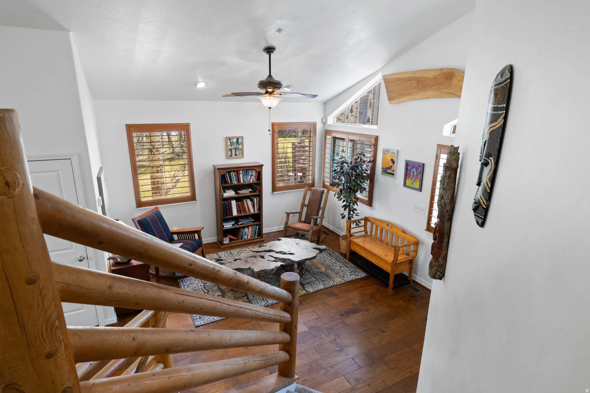 Vaulted ceiling living area featuring wood-finish flooring, multiple windows with wooden plantation shutters, and a ceiling fan with integrated lighting