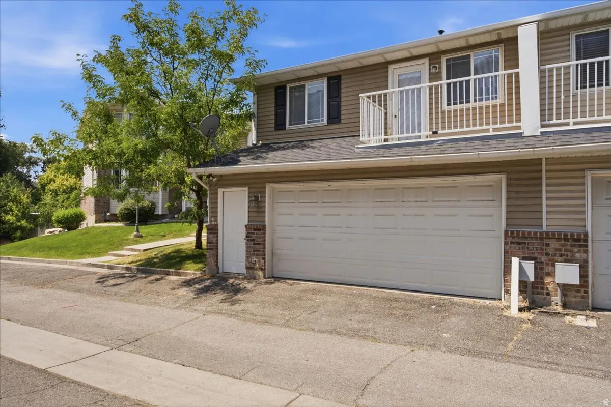 Two-car garage, upper-level balcony with white railing, siding exterior, brick accent, and mature landscaping