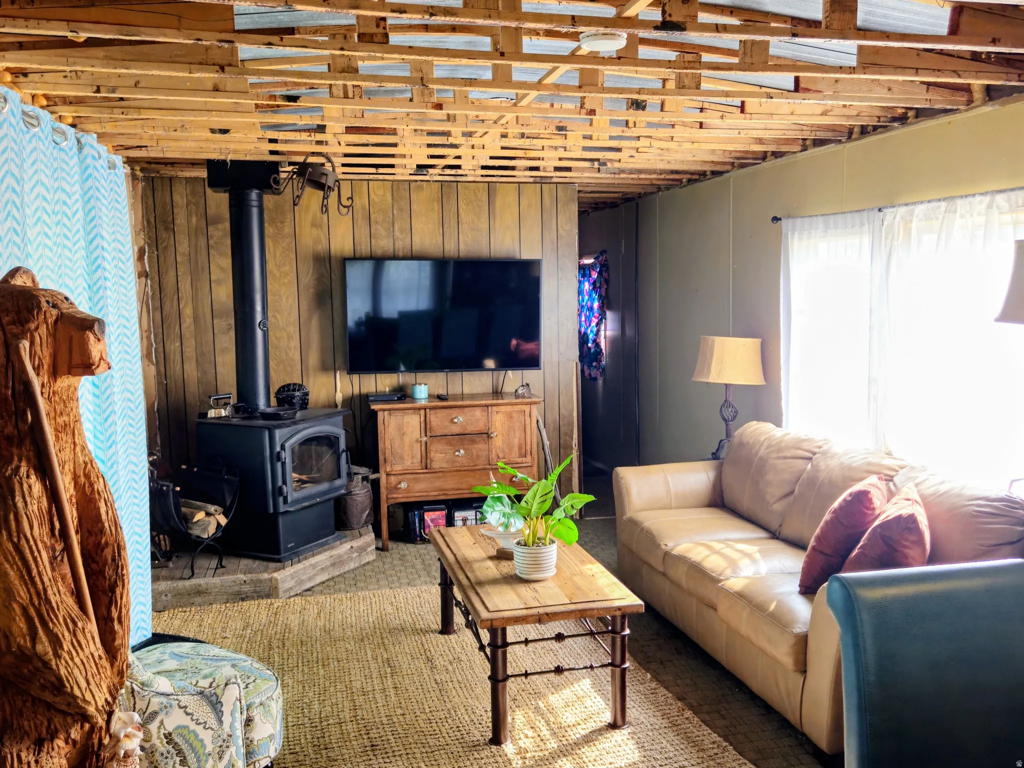 Wood-paneled wall accents, exposed wood-frame ceiling, black wood-burning stove on a raised hearth, large windows providing natural light, and textured area rug