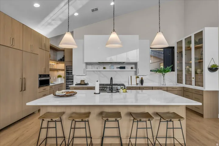 Spacious kitchen featuring a large central island with seating, integrated appliances, extensive light wood-finish cabinetry, a striking white range hood, and vaulted ceilings