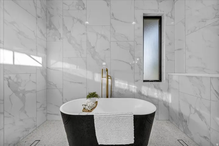 Freestanding black soaking tub, floor-mounted brass tub filler, white marble-look wall tiling, mosaic floor tiling, and a vertical frosted window