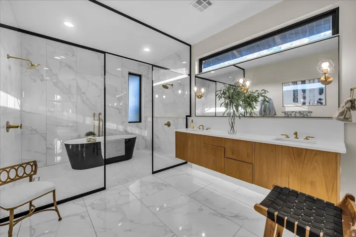 Floating wood-finish vanity with dual sinks, expansive wall-mounted mirror, white countertop, freestanding black bathtub, and glass-enclosed shower