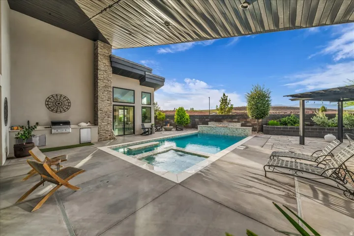 Covered patio with integrated outdoor kitchen, built-in swimming pool and spa, stone accent wall, and a slatted pergola