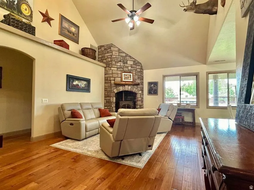 Vaulted ceiling with a ceiling fan, stone-clad fireplace with a wood mantle, wood-finish flooring, and multiple windows
