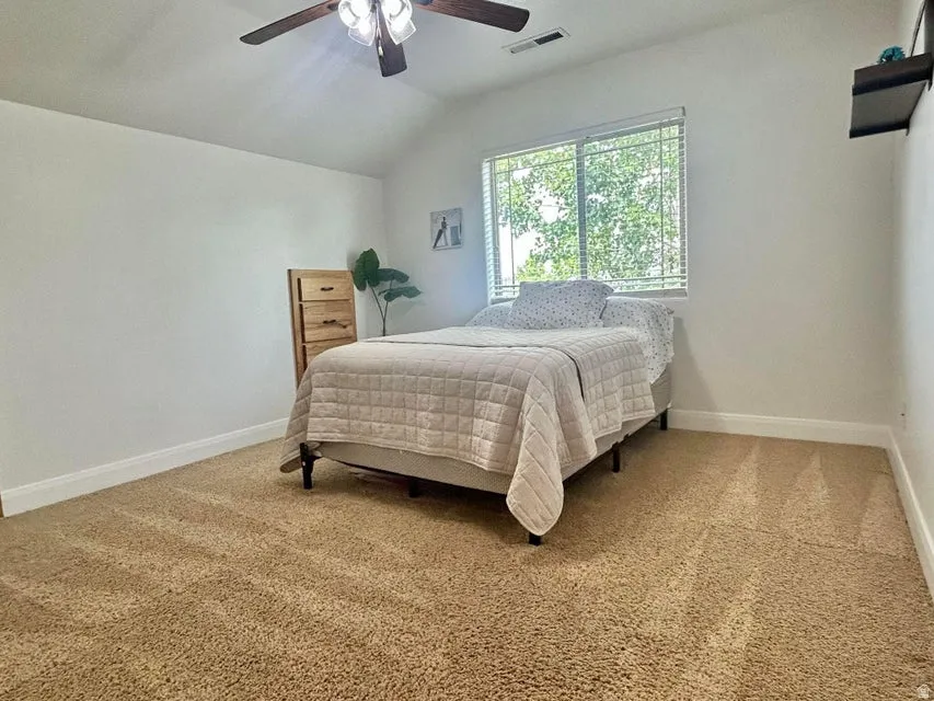 Vaulted ceiling, ceiling fan with integrated lighting, window with blinds, light-toned carpet, and a wall-mounted shelving unit