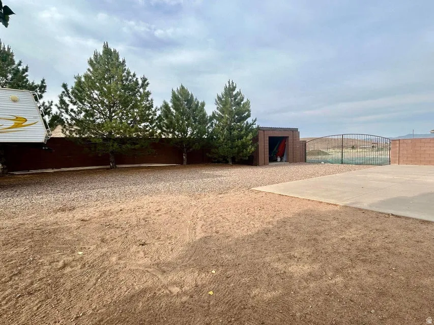 Gravel and dirt lot with mature evergreen trees, a concrete pad, a shed, and a metal fence