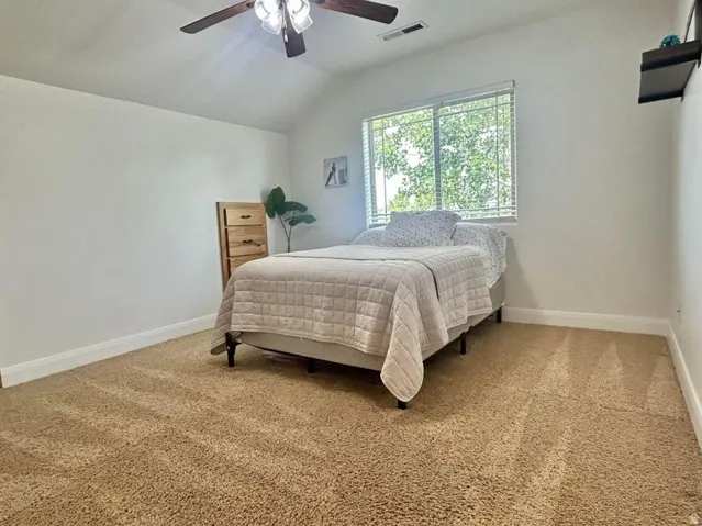 Vaulted ceiling, ceiling fan with integrated lighting, window with blinds, light-toned carpet, and a wall-mounted shelving unit