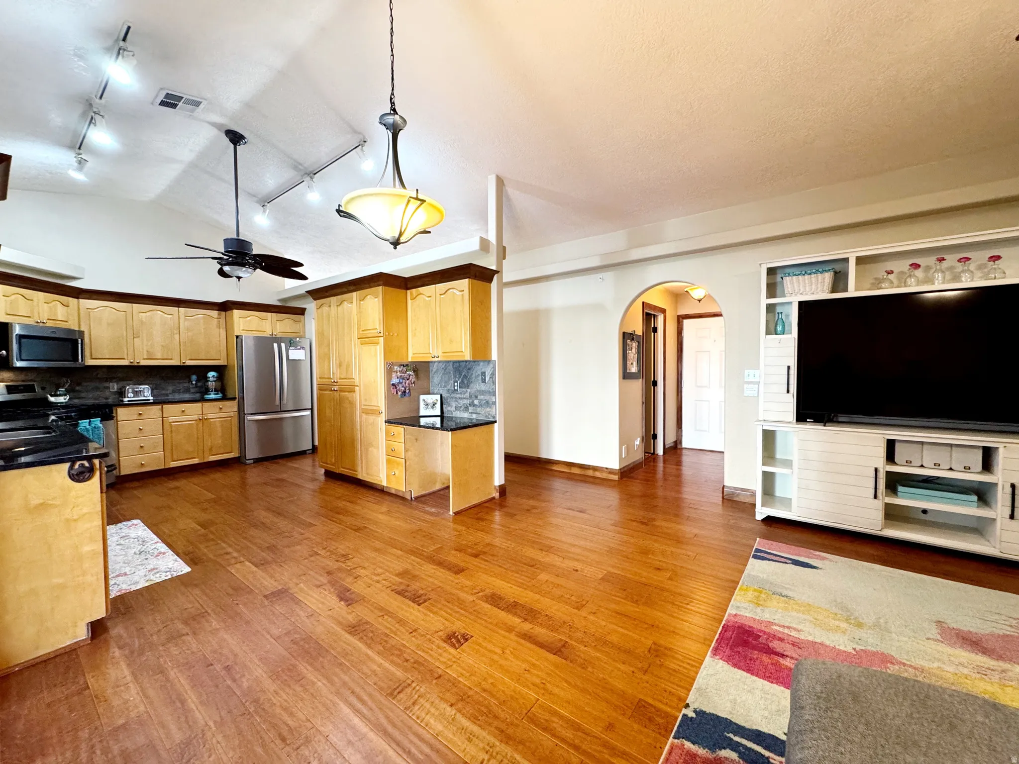 Vaulted ceiling with track lighting, wood-finish flooring, kitchen with light-toned cabinetry, built-in media console, and an arched doorway