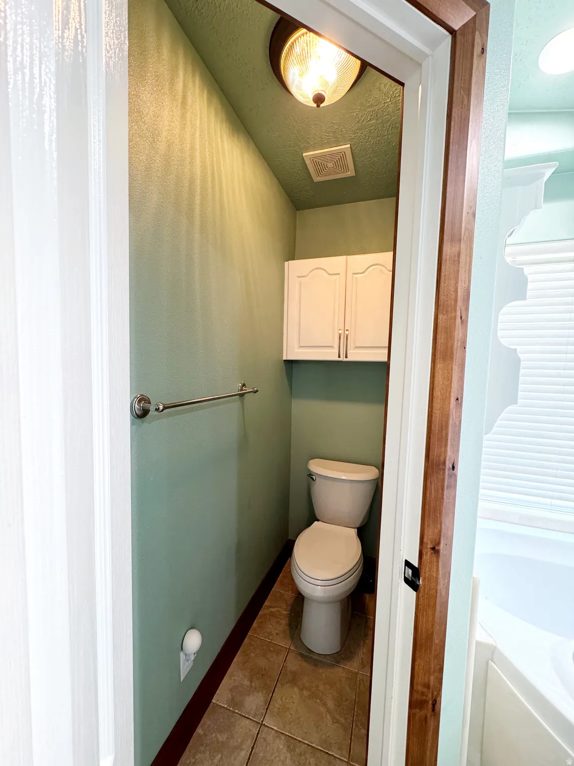 Compact water closet featuring a built-in toilet, tiled flooring, mounted wall cabinetry, and a towel bar