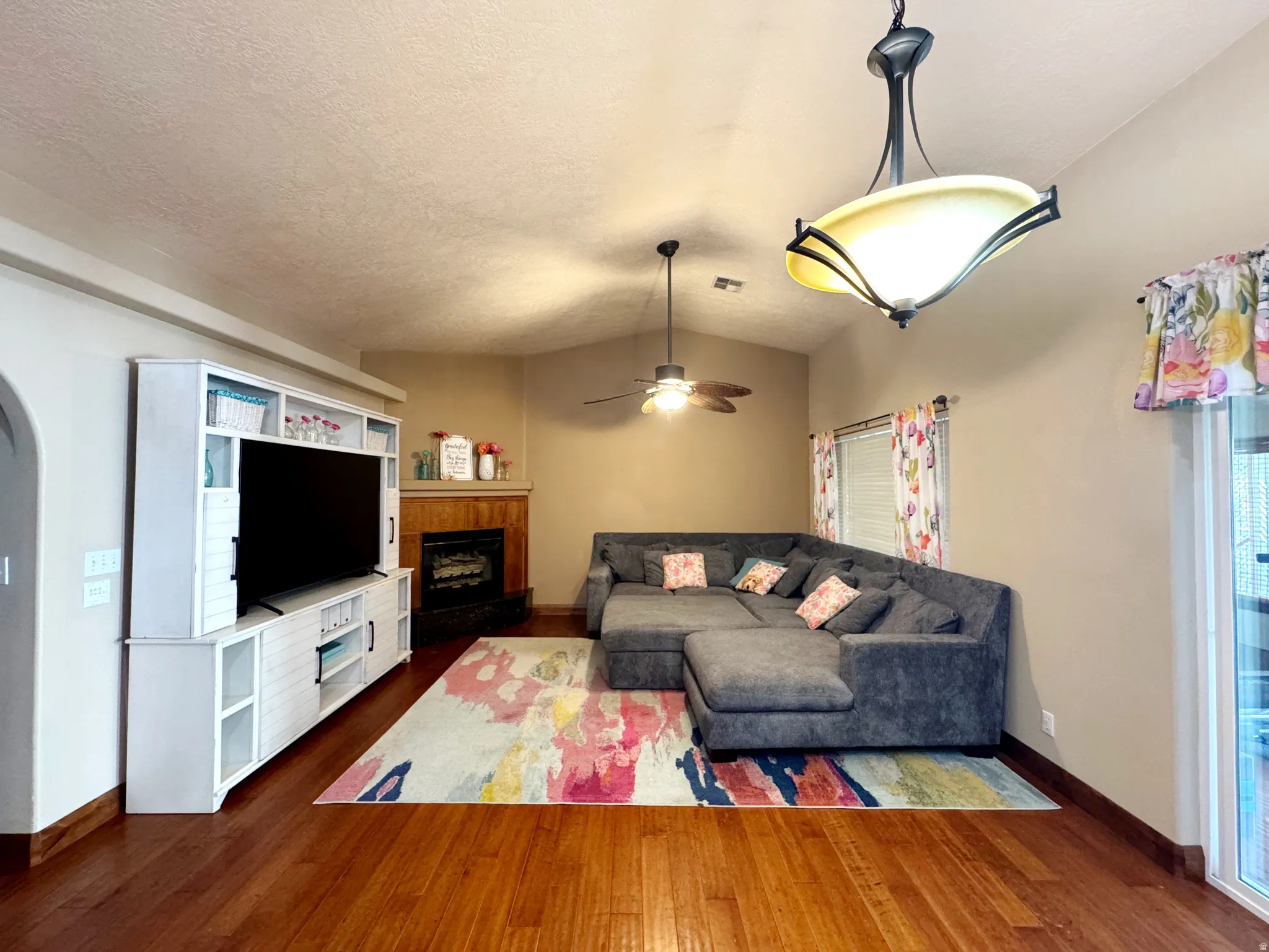 Vaulted ceiling, wood-finish flooring, built-in entertainment center, corner fireplace with wood mantel, and a ceiling fan