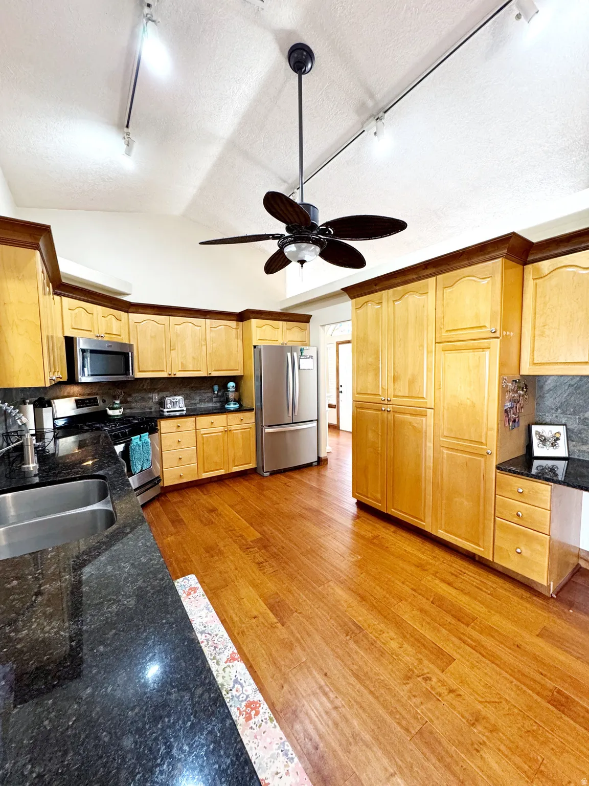 Kitchen featuring wood-finish flooring, light wood cabinetry, dark countertops, a double basin sink, and stainless steel appliances
