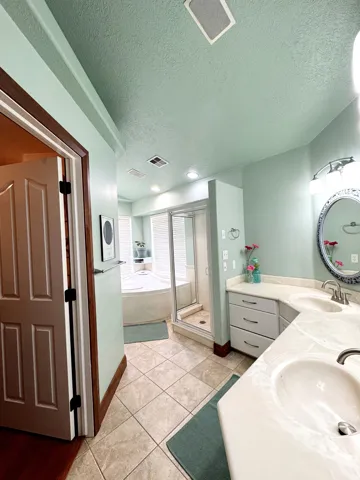 Dual vanity with integrated sinks, corner jetted tub, glass-enclosed shower, tile flooring, and recessed lighting