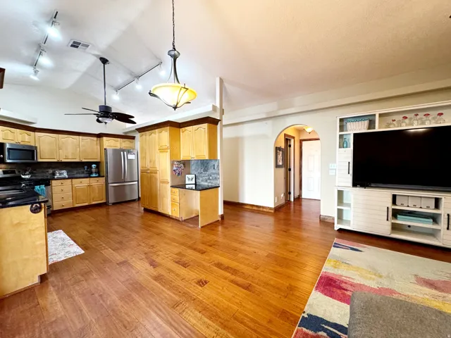 Vaulted ceiling with track lighting, wood-finish flooring, kitchen with light-toned cabinetry, built-in media console, and an arched doorway