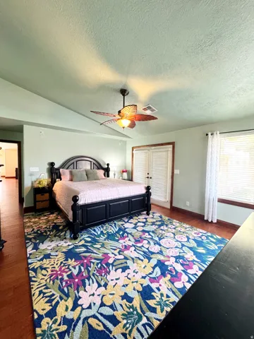 Vaulted ceiling, ceiling fan with integrated lighting, wood-finish flooring, white louvered double doors, and a window with blinds