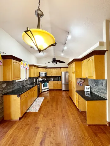 Vaulted ceiling kitchen featuring wood-finish flooring, light wood cabinetry, black countertops, and stainless steel appliances