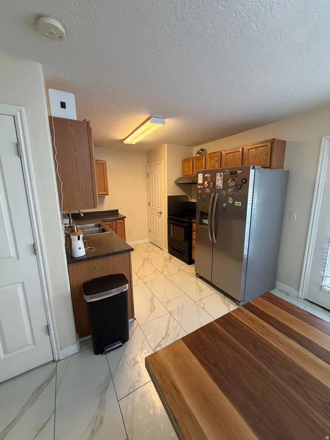 Kitchen featuring wood-finish cabinetry, stainless steel refrigerator, black range, black countertops, and light-toned tile flooring