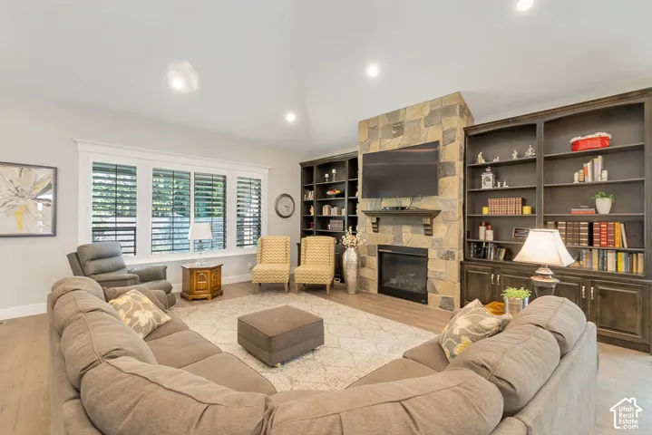Stone-clad fireplace with mantel, built-in shelving, triple window with plantation shutters, recessed lighting, and wood-finish flooring