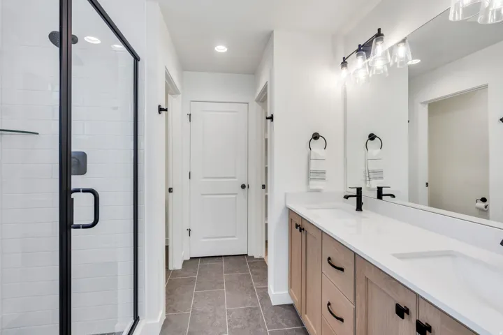 Walk-in shower with black-framed glass enclosure, double vanity with light wood-finish cabinetry, dual undermount sinks, and white countertops