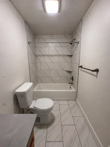 Bathtub with tile surround and built-in shelving, white toilet, and tile flooring