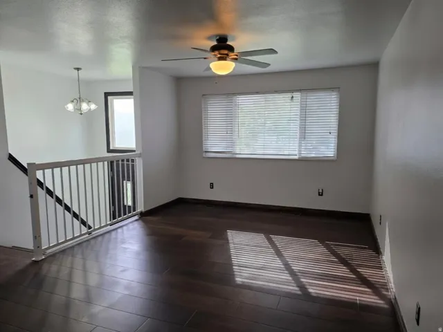 Spacious open-concept living area featuring dark wood-finish flooring, abundant natural light from expansive multi-pane windows, ceiling fan with integrated lighting, and a railed staircase