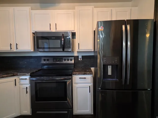 Modern kitchen featuring white cabinetry with black hardware, black stainless steel appliances including a French-door refrigerator and electric range, dark tile backsplash, and stainless steel microwave mounted above the cooktop